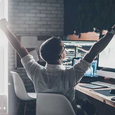 Office worker raising both arms in front of trading screens